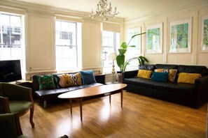 A spacious living room with a wooden floor, a black leather sofa with colorful cushions, a wooden coffee table, and a large window providing natural light. - Cozy 3-Bedroom Flat - Fitzrovia & Soho (London)