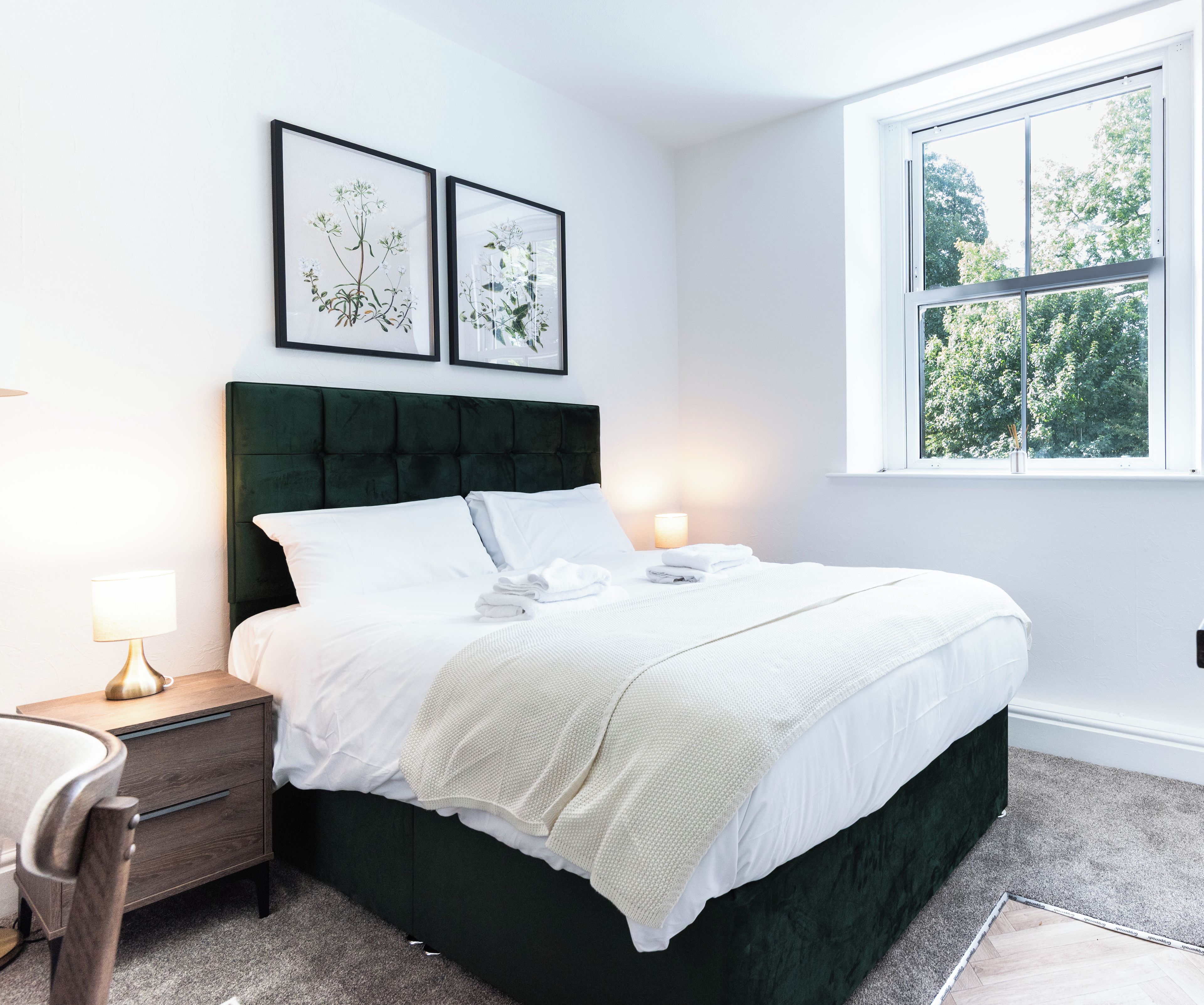 A bedroom with a bed, bedside tables, a window, and framed botanical prints on the wall.