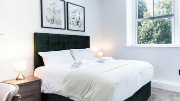 A bedroom with a bed, bedside tables, a window, and framed botanical prints on the wall.