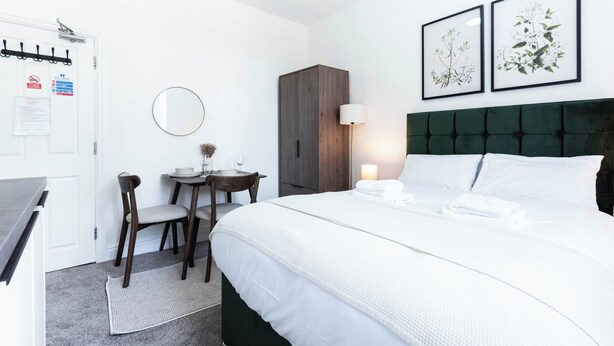 Quarto de hotel com cama, escrivaninha com cadeira, guarda-roupa, espelho redondo e dois quadros com gravuras de plantas na parede.