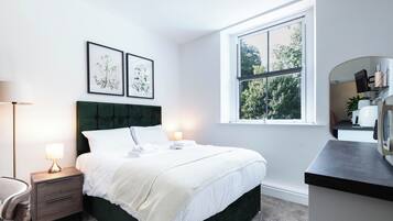 A hotel room with a bed, bedside table, lamp, framed botanical prints, a window with a view of trees, and a mirror.