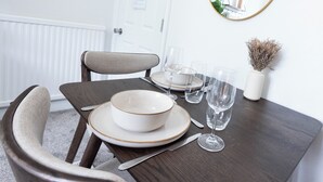 Uma mesa de jantar posta com pratos, tigelas, talheres e copos de vinho, com uma cadeira e um vaso com flores secas ao lado.