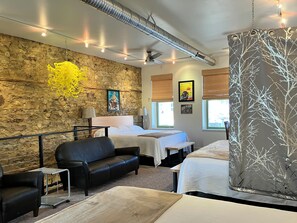 Habitación de hotel con pared de piedra, una cama, un sofá, una mesita y una ventana con persianas.