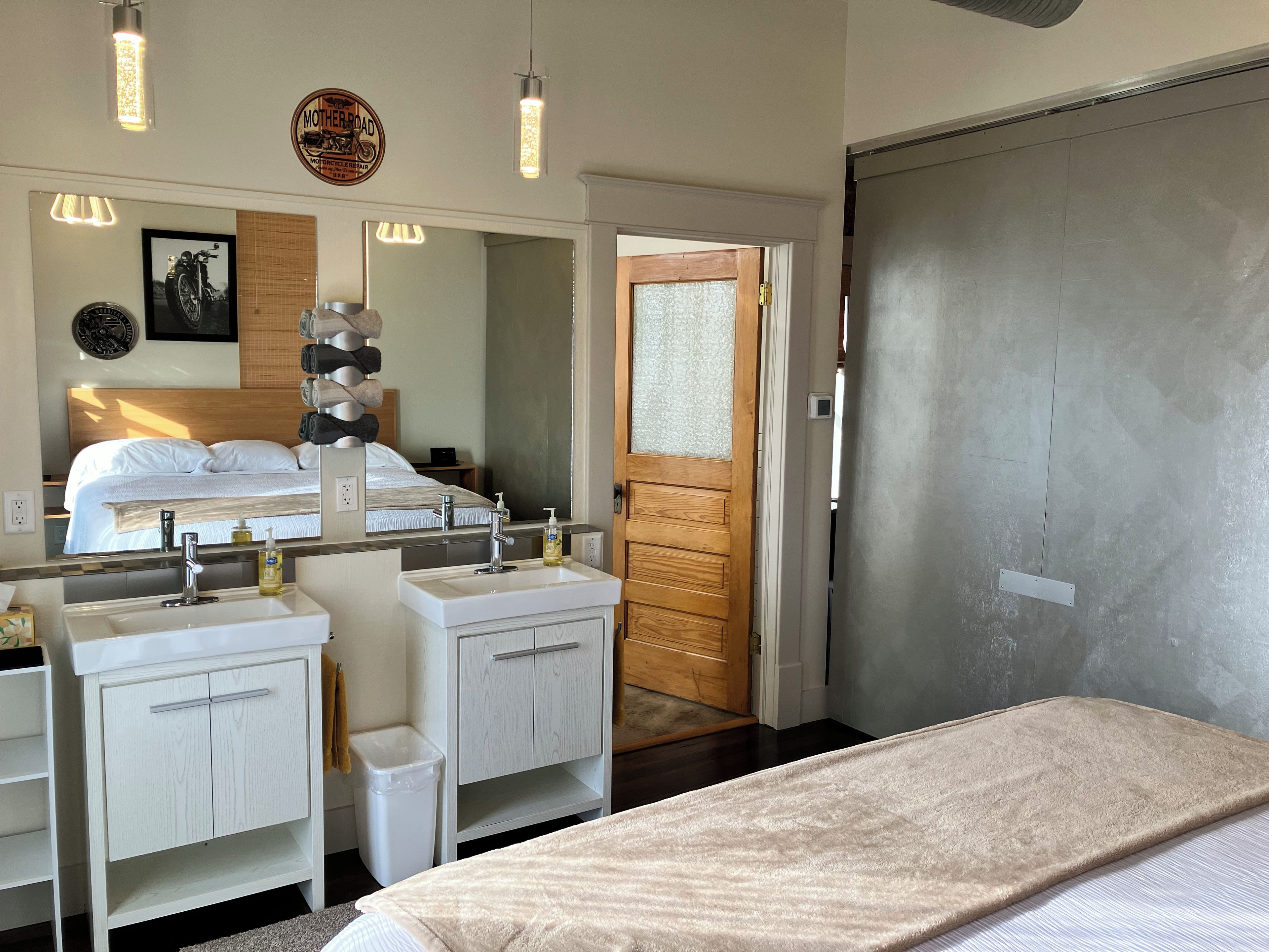 A hotel room with a bed, two sinks, a mirror, a wooden door, and a wall-mounted light fixture.