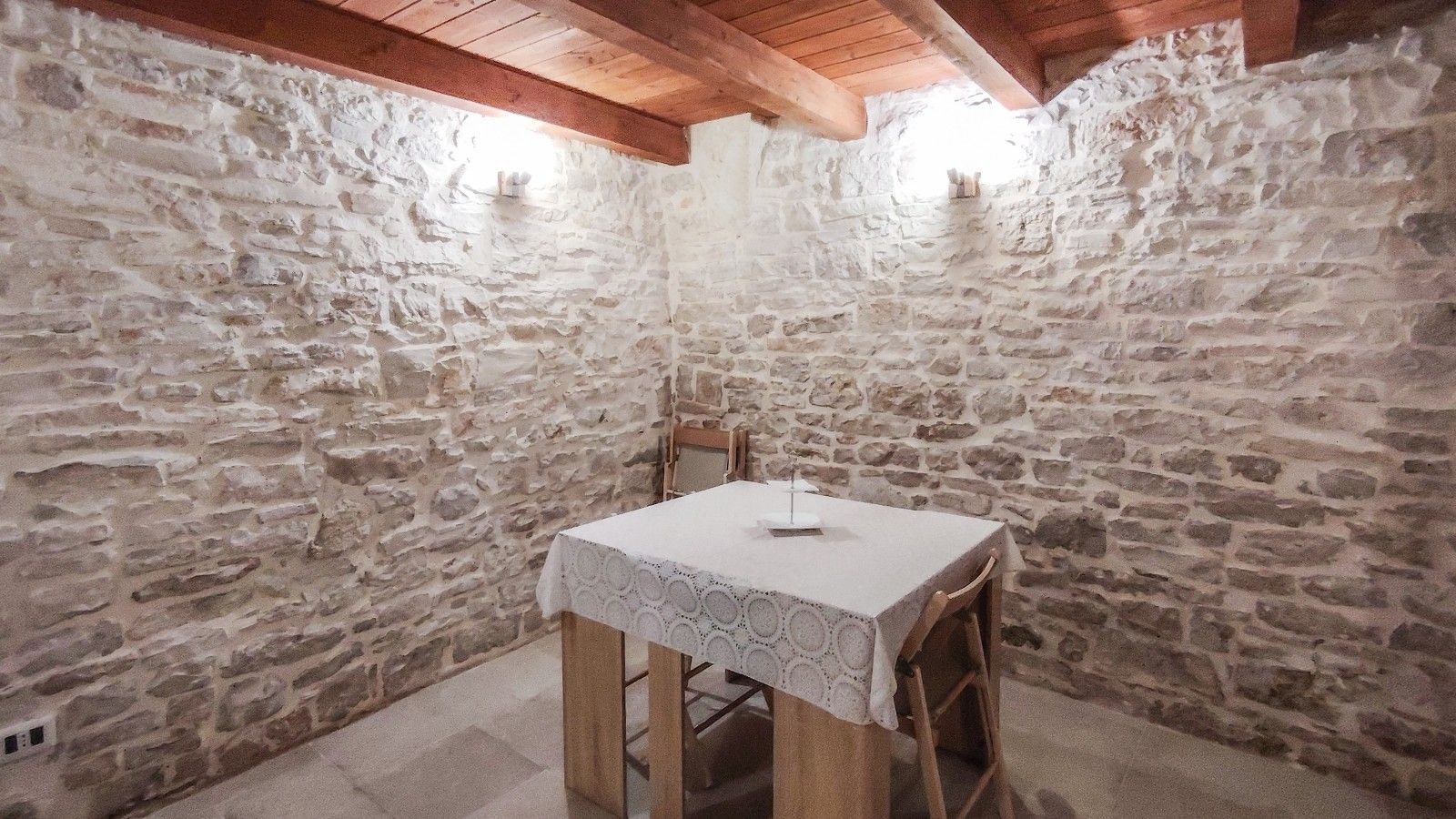 A stone-walled room with a table and chairs, wooden beams on the ceiling, and a small window.