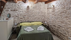 Una habitación con pared de piedra, cama con ropa de cama verde, mesita de noche con lámpara y una pequeña mesa con una cesta y un vaso.