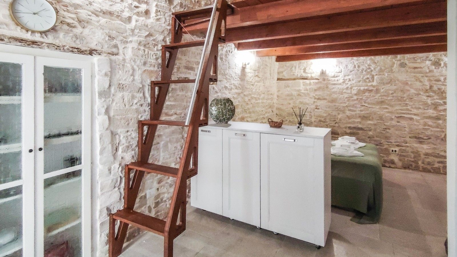 Un intérieur avec un mur en pierre, une échelle en bois, des armoires blanches et une table verte.