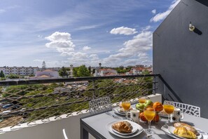 Ein Balkon mit Frühstückstisch, auf dem Croissants, Eier und Orangensaft bereitstehen, mit Blick auf eine Stadtlandschaft.