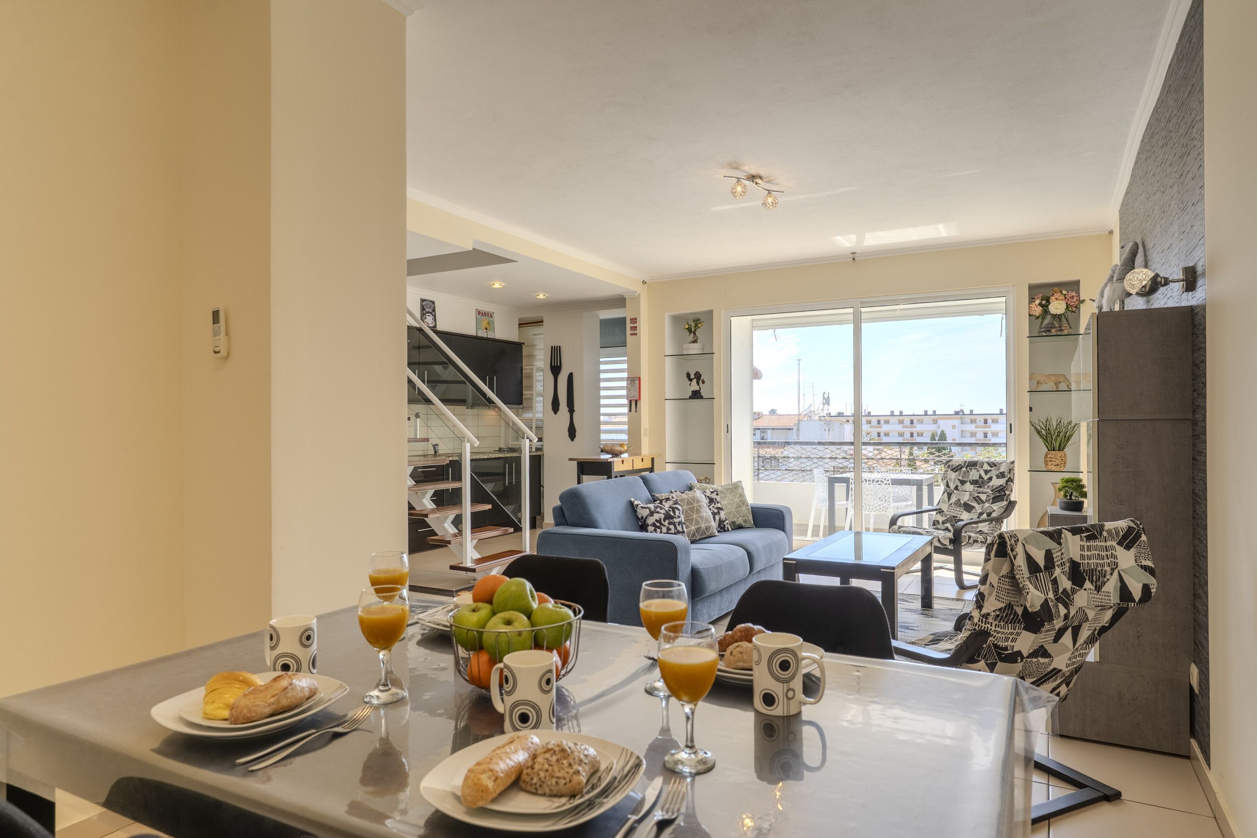 A dining area with a table set for breakfast, a blue sofa, and a view of the city through large windows.