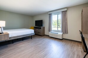 Una habitación de hotel con cama, televisor, escritorio con lámpara, ventana con cortinas y aire acondicionado integrado.