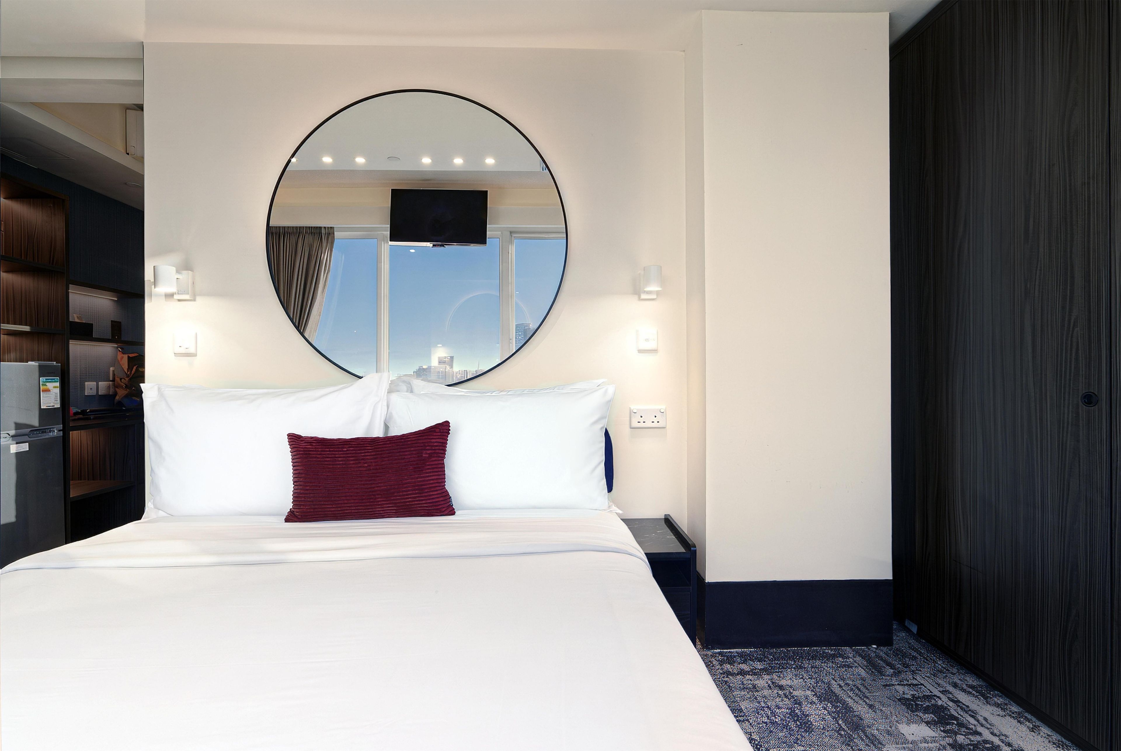 A hotel room with a large bed, a round mirror above it, and a view of the city through the window.