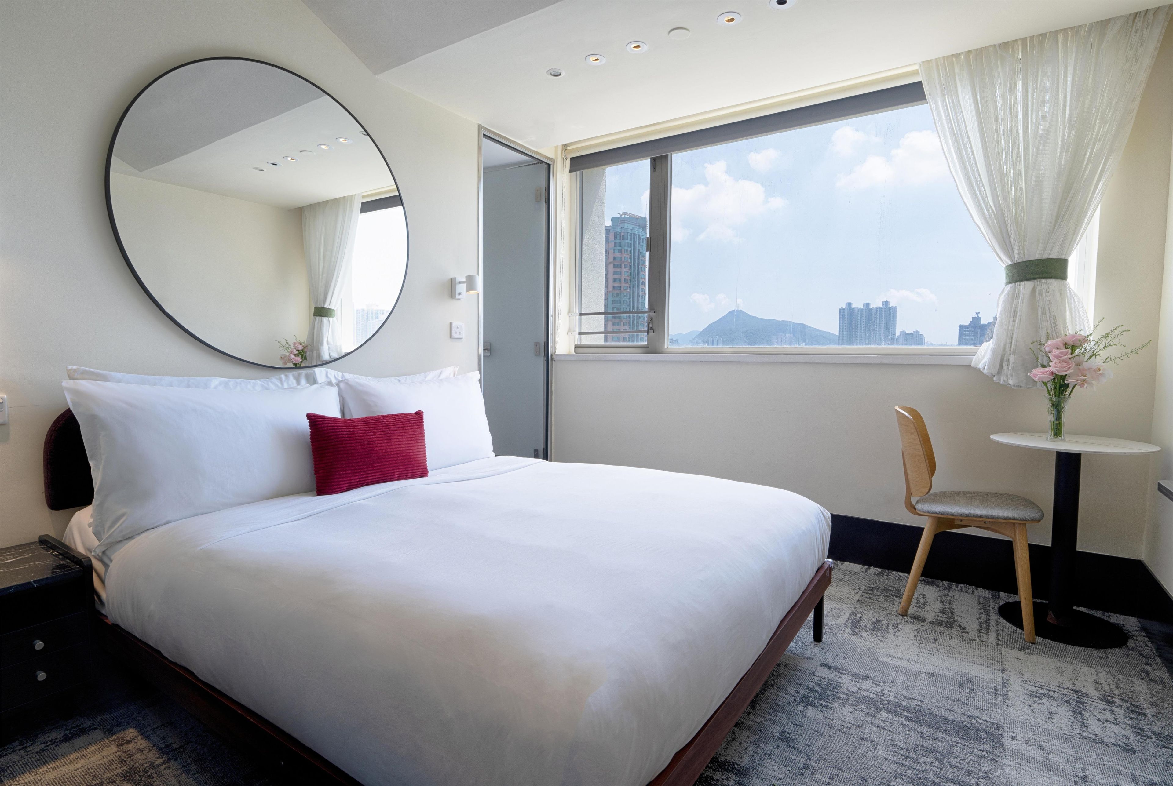 Habitación de hotel con una cama grande, un espejo redondo, una ventana con vistas a edificios, una mesita con una silla y un jarrón con flores.