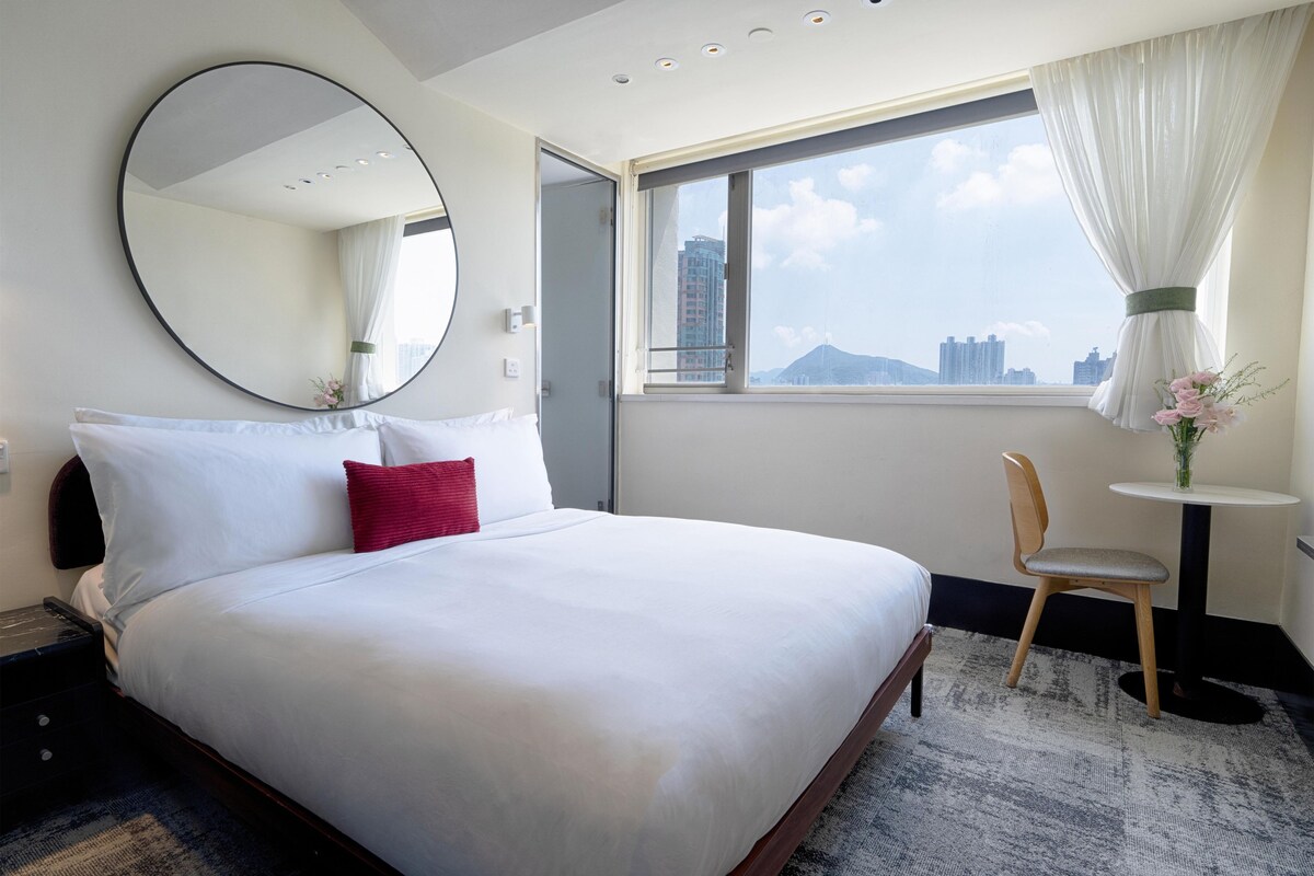 Une chambre d’hôtel avec un grand lit, un miroir rond, une fenêtre donnant sur des immeubles, une petite table avec une chaise et un vase rempli de fleurs.