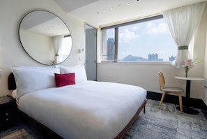 Quarto de hotel com uma cama grande, um espelho redondo, janela com vista para edifícios, uma mesa pequena com uma cadeira e um vaso com flores.