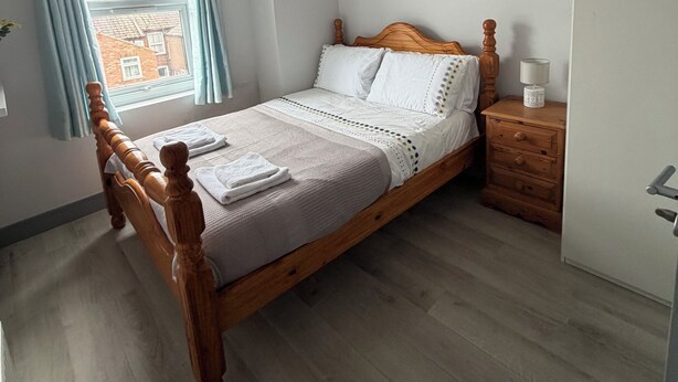 Quarto com cama de madeira, mesa de cabeceira com abajur e janela com cortinas.
