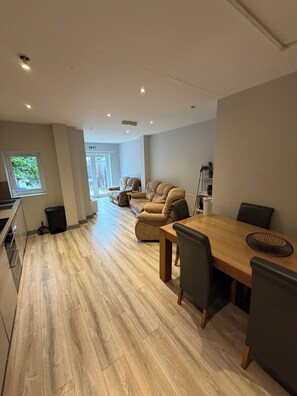 A modern kitchen with wooden flooring, a dining area with a wooden table and chairs, and a seating area with a sofa and armchairs. - Zana House - 6 Bedroom Contractor, Families and Group Accomodation (Ipswich)
