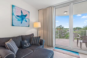 Een moderne woonkamer met een donkergrijze bank, decoratieve kussens, een schilderij van een zeester, een vloerlamp en uitzicht op een balkon met buitenmeubilair.