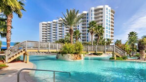 A swimming pool with a wooden bridge, surrounded by palm trees and a multi-story building in the background. - 🌴Beach Nearby + Lagoon Pool | Fab Amenities | Palms Resort 1404☀️ (Destin)