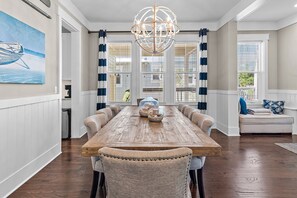 A dining room with a wooden table, upholstered chairs, a chandelier, and large windows with blinds. - 🌴Sea Esta | 4BR Coastal Retreat | Golf Cart + 6 Bikes | NatureWalk🌴 (Santa Rosa Beach)