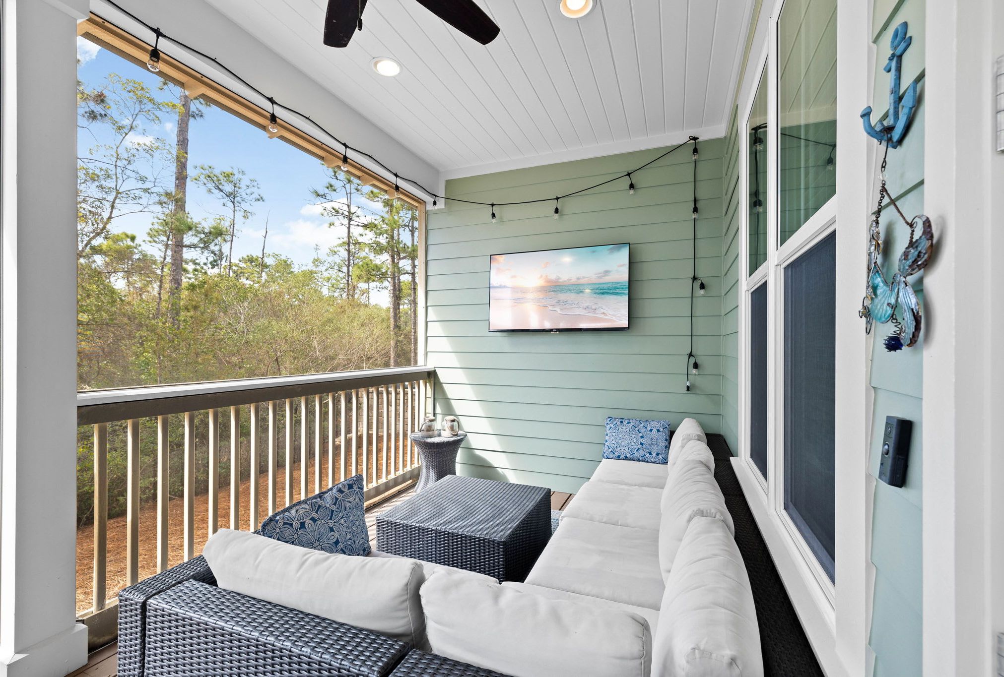 A covered porch with a view of trees and a beach, furnished with a sofa and a table.