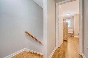 Un pasillo con escalera de madera y una puerta que da a una habitación con ventana y una silla.