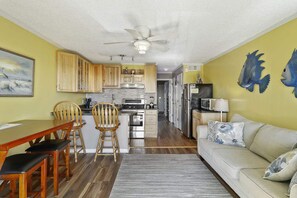 A compact kitchen with wooden cabinets, a dining area with a table and chairs, a ceiling fan, and a living area with a sofa and decorative pillows.