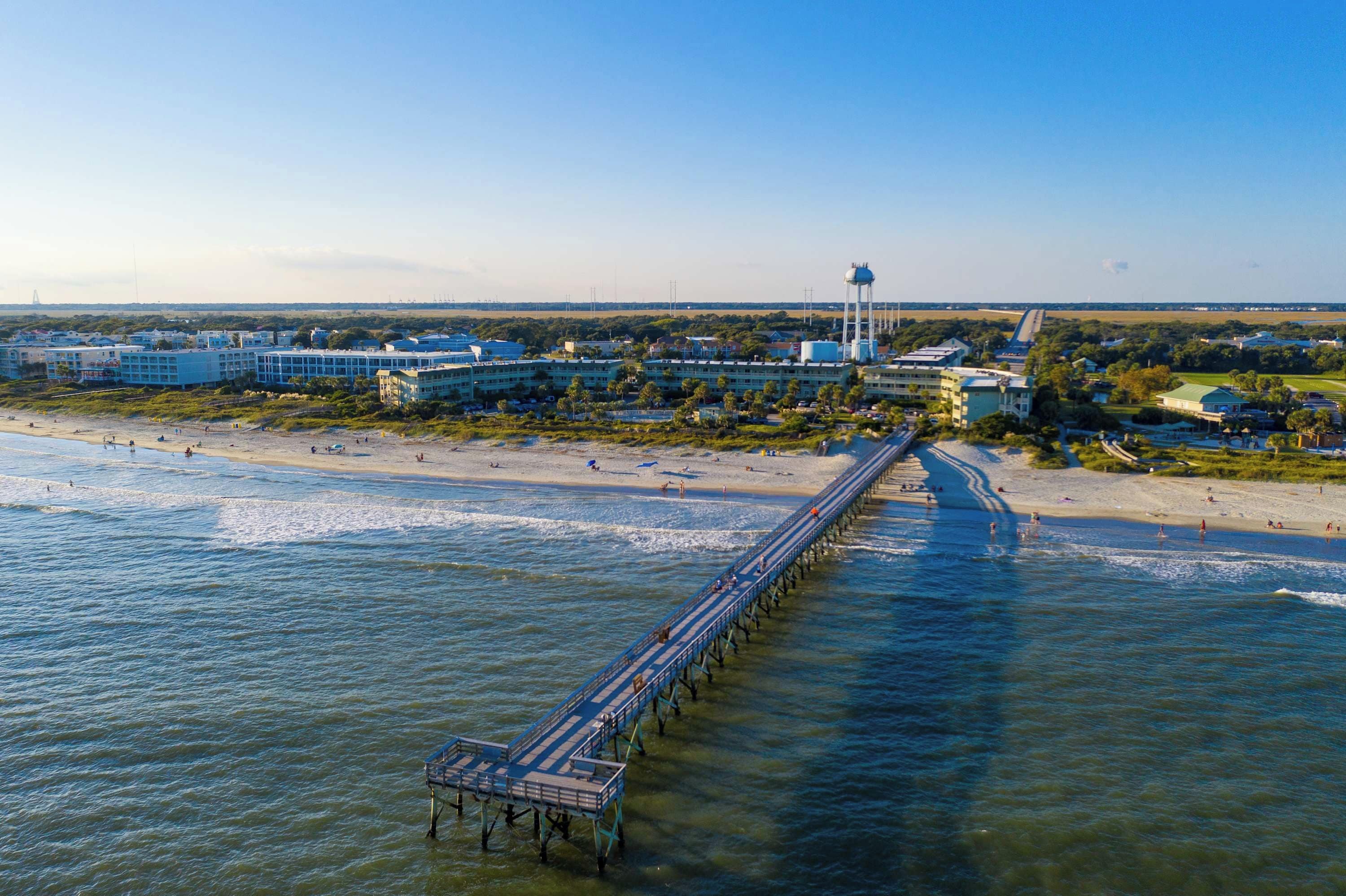 Una zona costera con un muelle largo que se extiende sobre el agua, una playa y edificios al fondo.