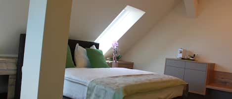Quarto com teto inclinado, cama com colcha estampada, mesa de cabeceira com abajur e cômoda.