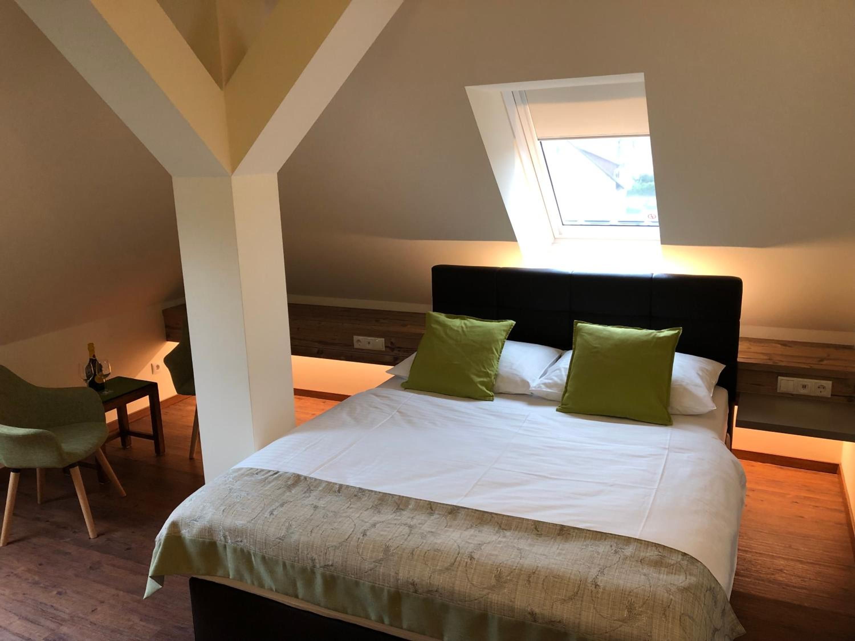 Ein Schlafzimmer mit einem Bett, grünen Kissen, einem Holzkopfteil, einem kleinen Tisch mit einer Flasche und einem Stuhl.