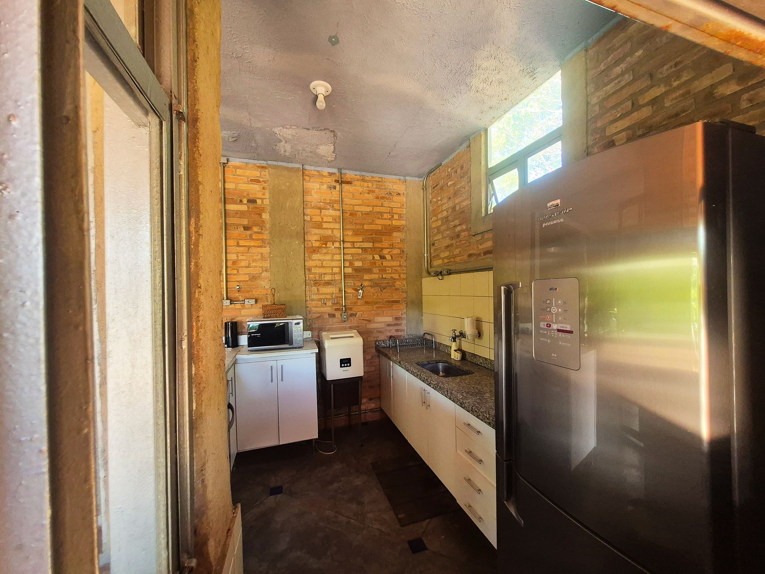 Cocina compacta con refrigerador de acero inoxidable, microondas y tostadora. Paredes de ladrillo visto y ventana que proporciona luz natural.