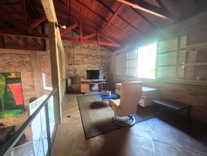 A room with a brick wall, a wooden desk, a television, a chair, and a window with blinds.