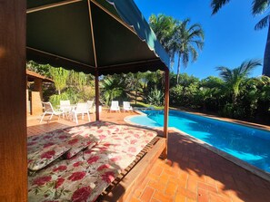 A pool area with a covered seating area, a pool, and surrounding greenery.
