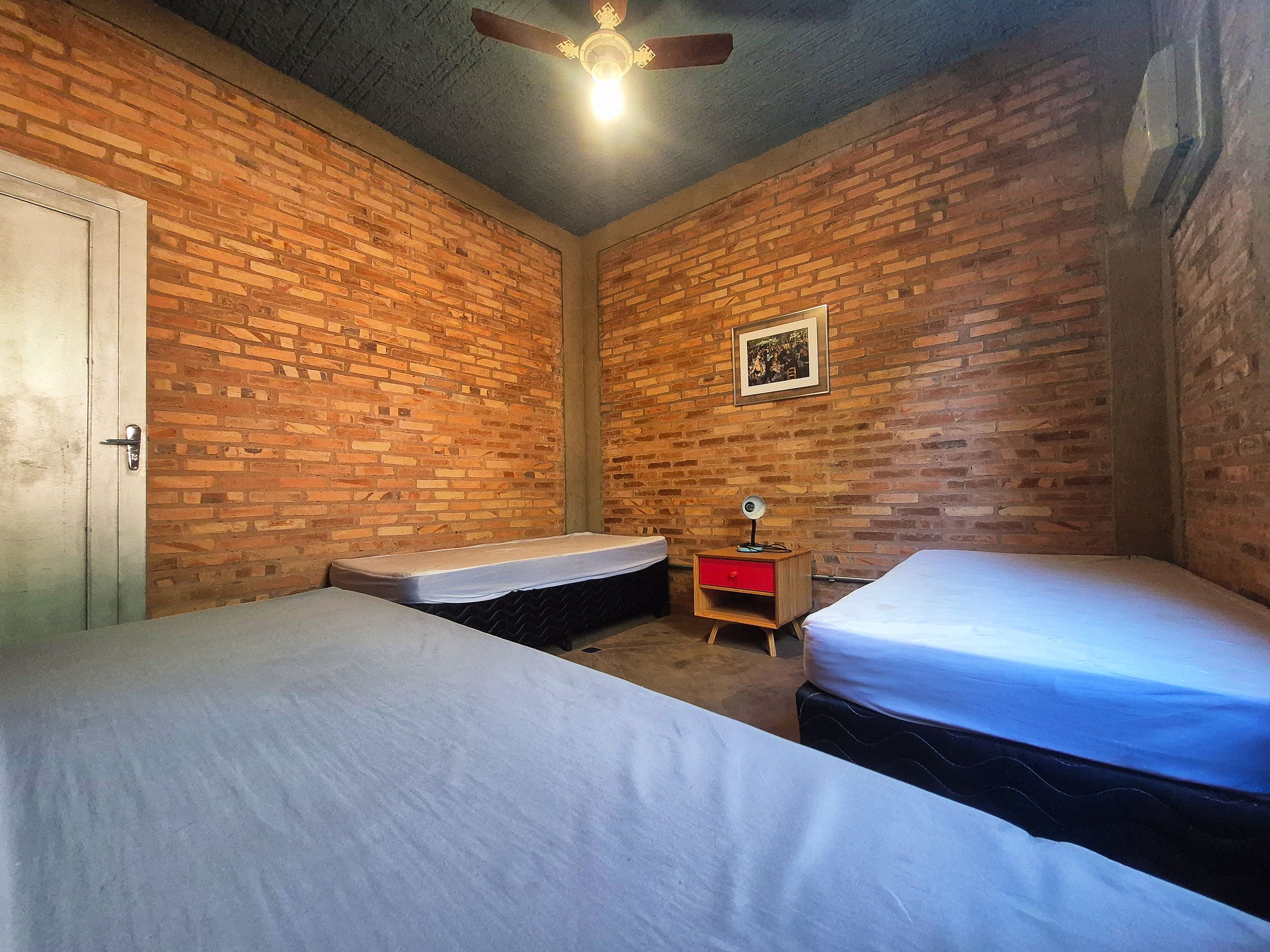 A room with two beds, a wooden nightstand with a red drawer, a ceiling fan, and a brick wall.