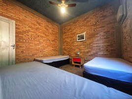 Quarto com duas camas, uma mesa de cabeceira de madeira com uma gaveta vermelha, um ventilador de teto e uma parede de tijolos.