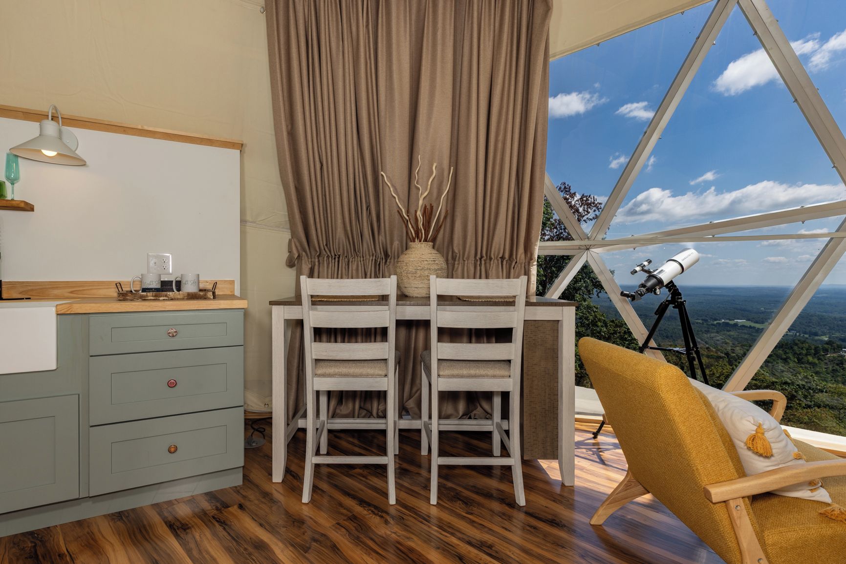 A room with a dining table, chairs, a telescope, and a view of the landscape through a large window.
