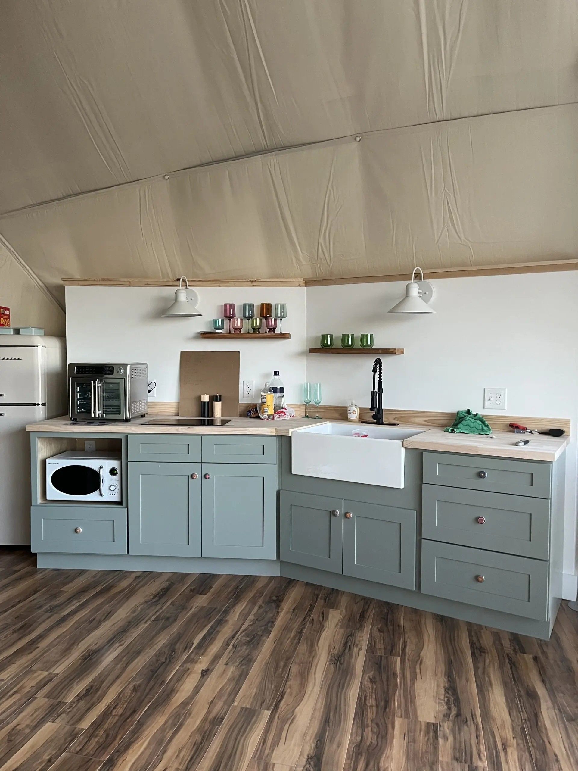 Cocina compacta con microondas, tostadora y lavamanos. Hay estantes de madera sobre la encimera y un techo abovedado.