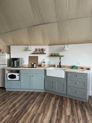 Een compacte keuken met een magnetron, broodrooster en een spoelbak. Boven het aanrecht zijn houten planken en het plafond is met stof bedekt.