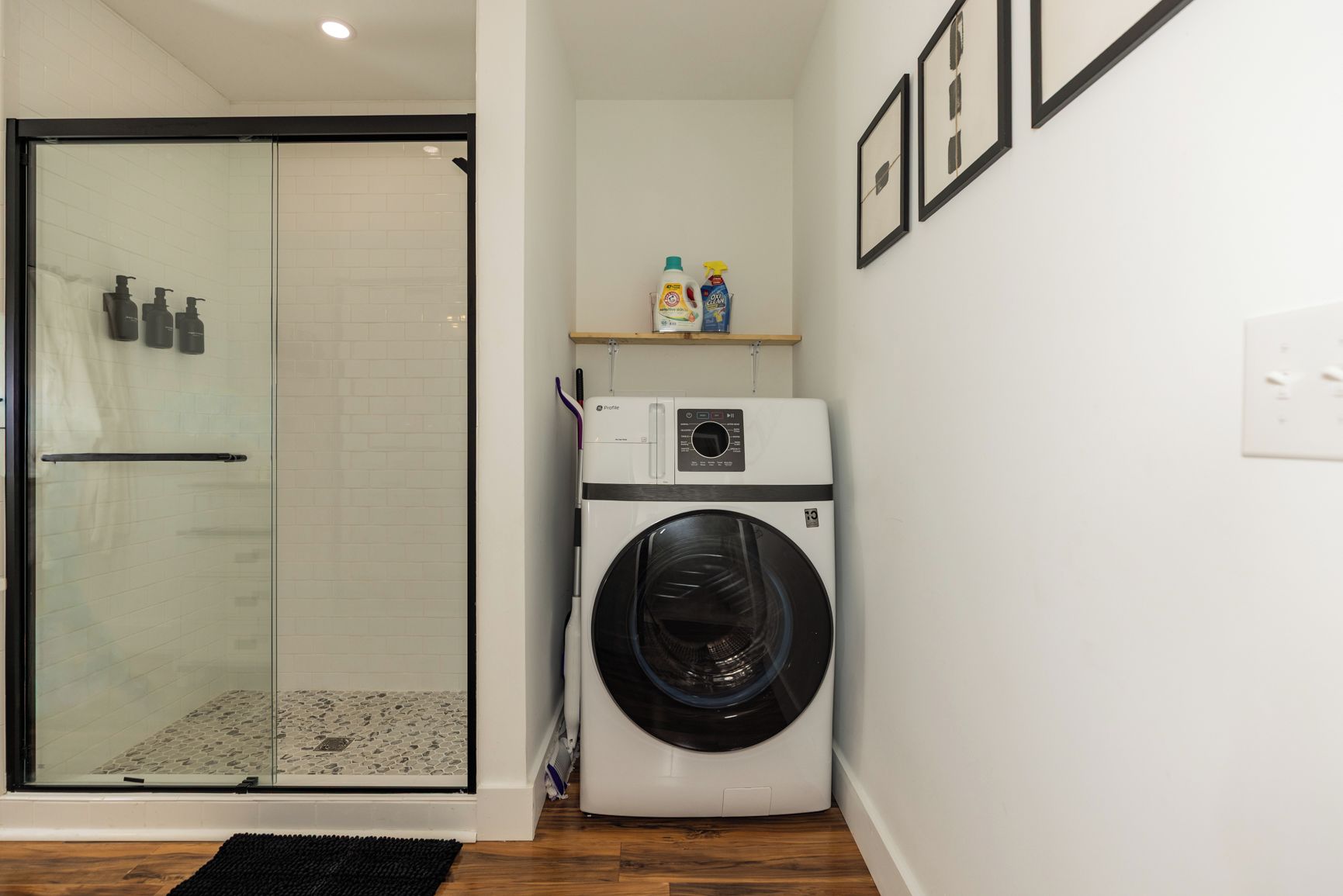 A compact laundry room with a glass shower enclosure, a washing machine, and a shelf with cleaning products.