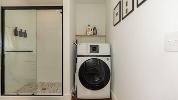 A compact laundry room with a glass shower enclosure, a washing machine, and a shelf with cleaning products.
