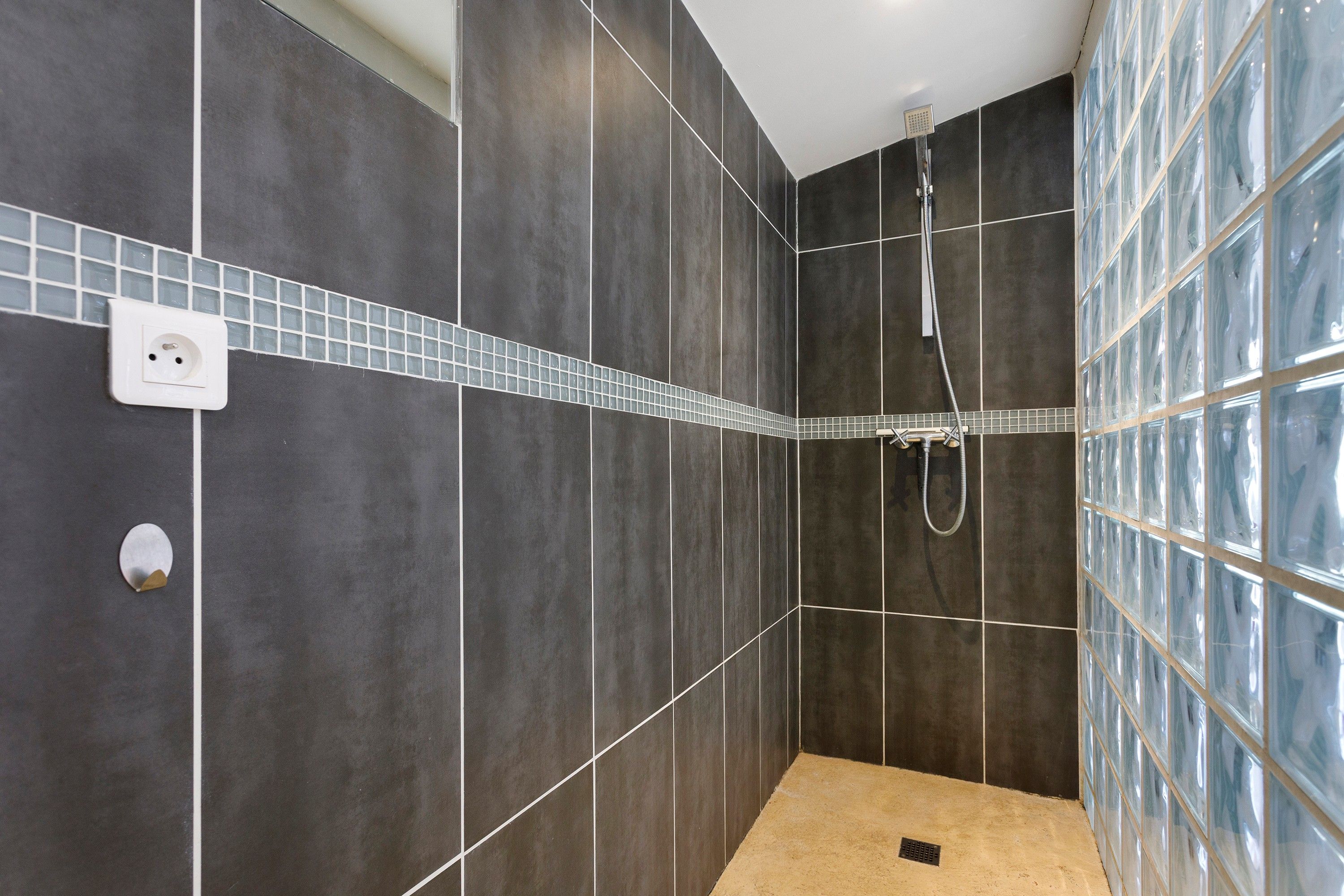 A modern bathroom with dark tiles, a glass shower enclosure, and a visible electrical outlet.