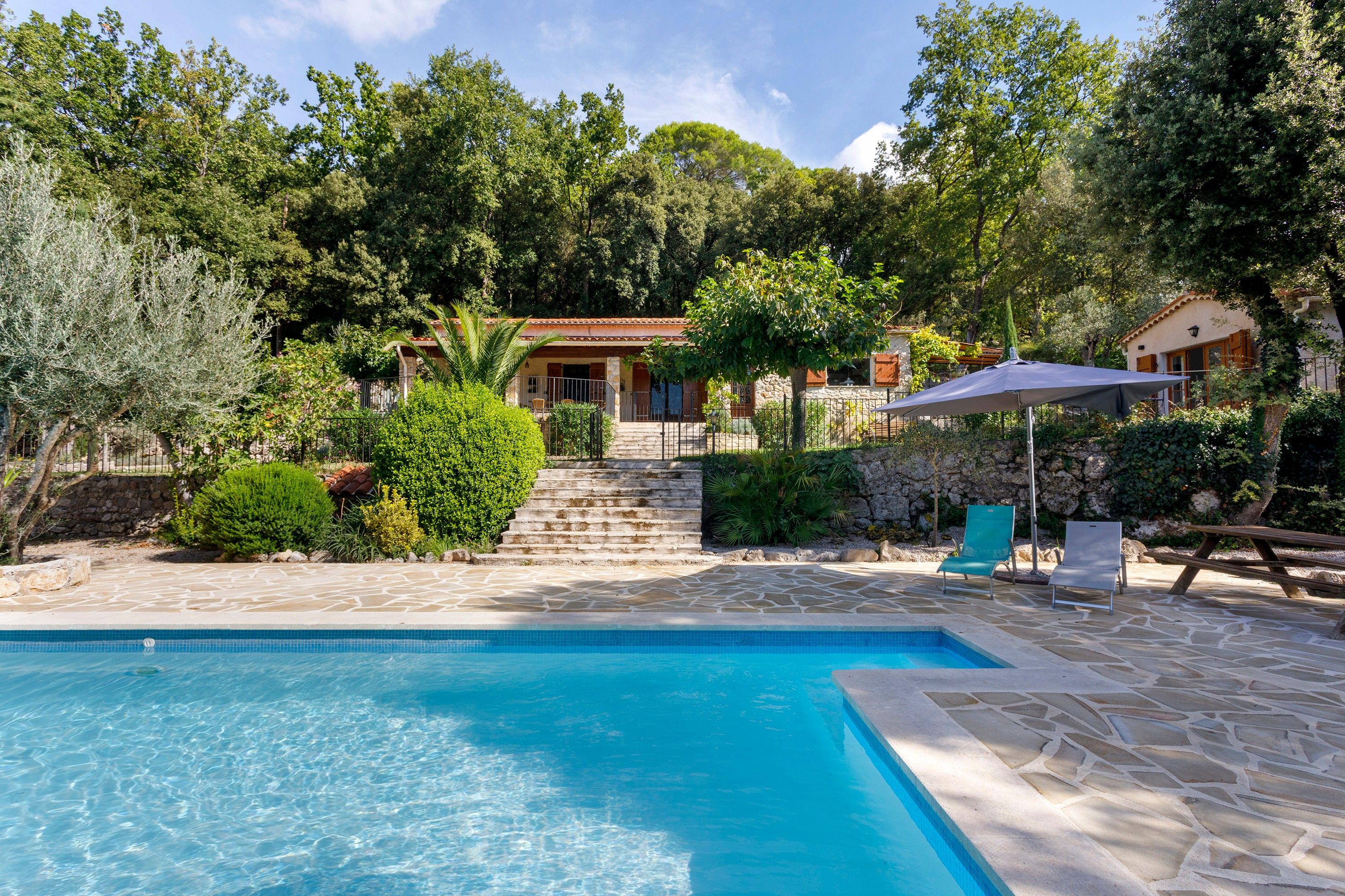 Un espace piscine doté d’une terrasse en pierre, d’une piscine à l’eau d’un bleu limpide et, au fond, d’un bâtiment avec un toit recouvert de tuiles.