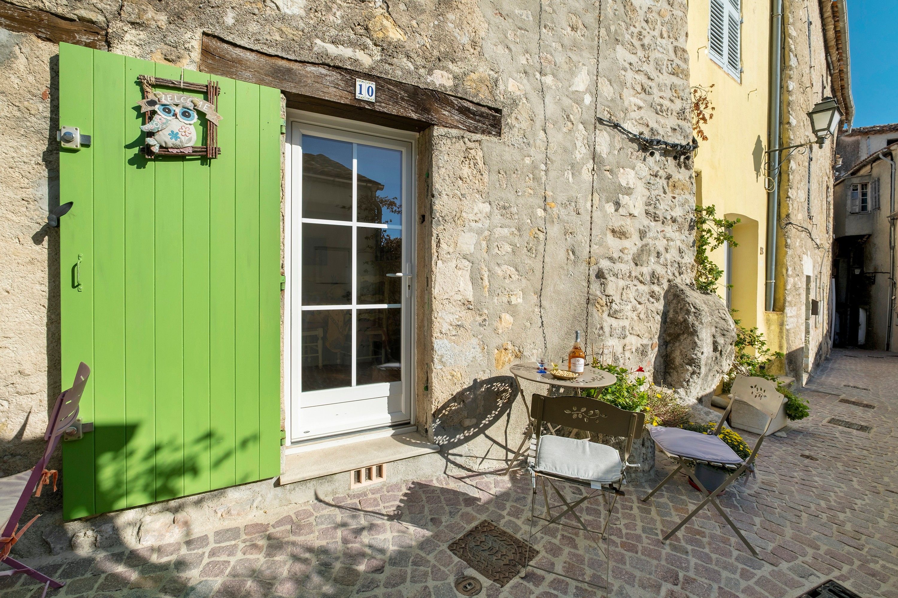 A stone building with a green door, a small outdoor seating area with a table and chairs, and a number '10' sign above the door.