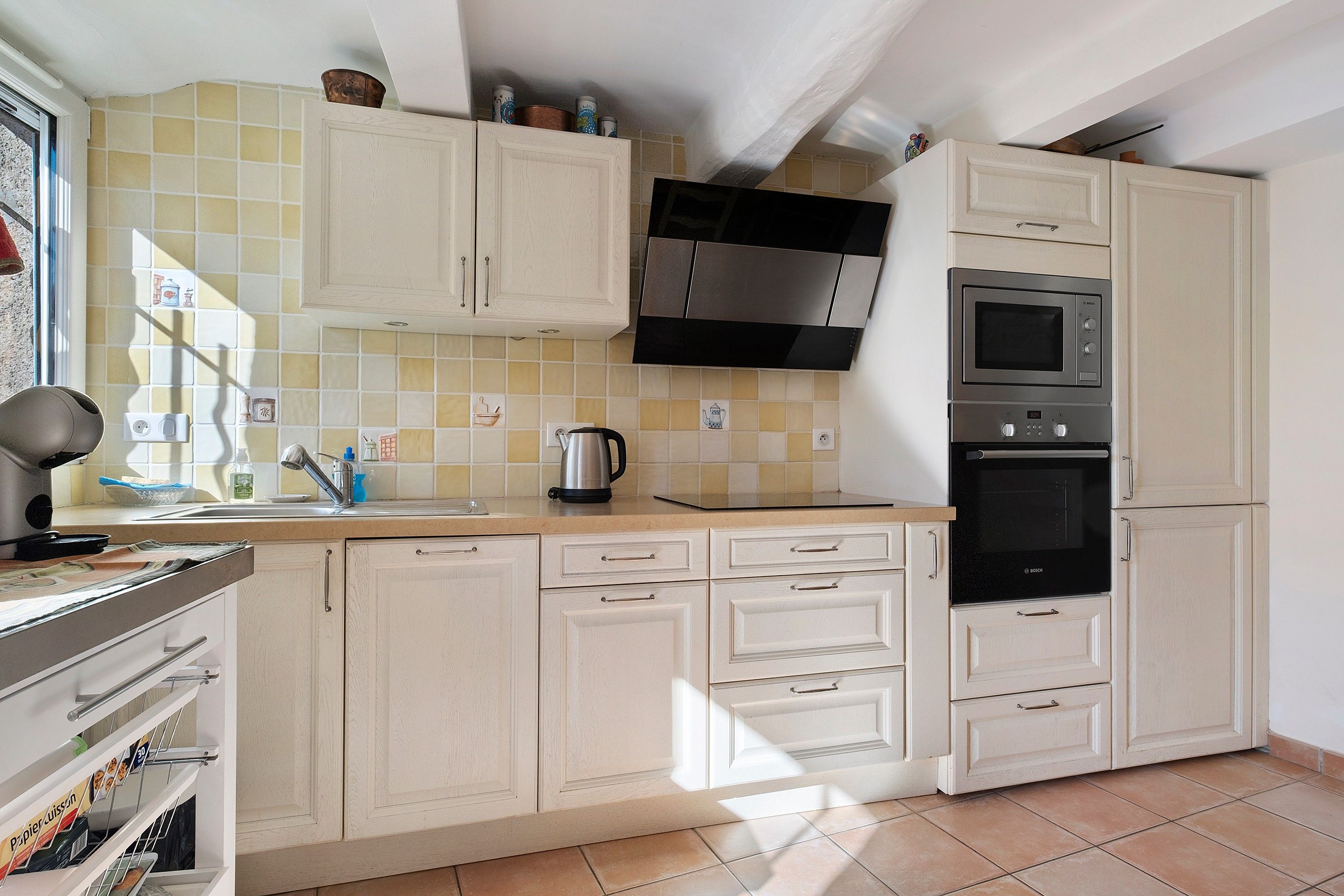 Una cocina con armarios blancos, una campana extractora de acero inoxidable, un horno Bosch y una tetera sobre la encimera.