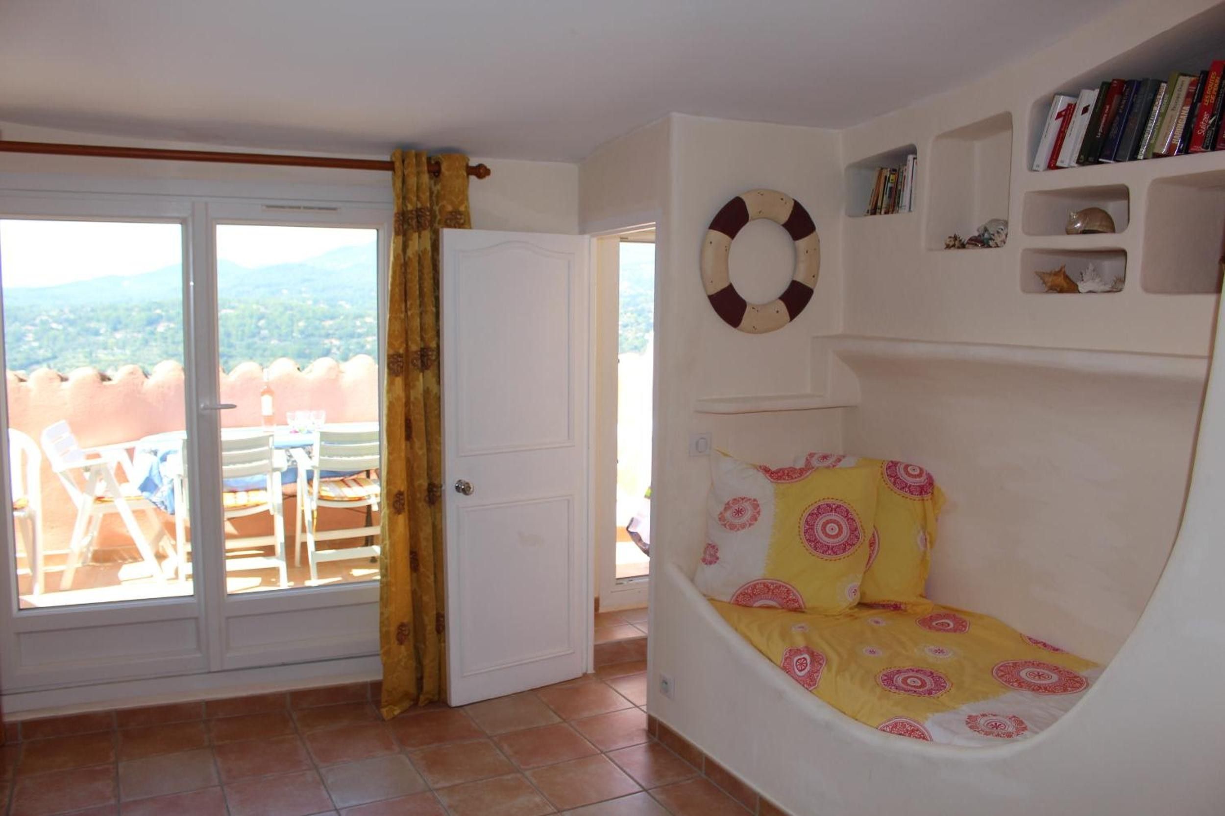 A room with a white door, a view of the outdoors, a white wall with a shelf, and a yellow and pink patterned cushion.