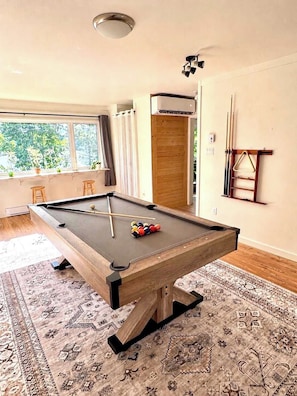 A pool table in a room with a wooden floor, a window with curtains, and a door.