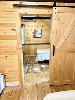 Ein Zimmer mit Holzboden, einem Bett, einem Gemälde an der Wand und einer Tür zu einem anderen Raum.