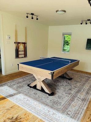 A pool table in a room with a patterned rug, a window with a view of greenery, and a ceiling light fixture.