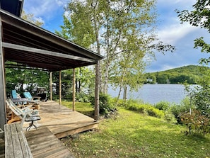 Eine Holzterrasse mit Liegestühlen und einem Grill, mit Blick auf einen See, in dessen Ferne ein Haus liegt.