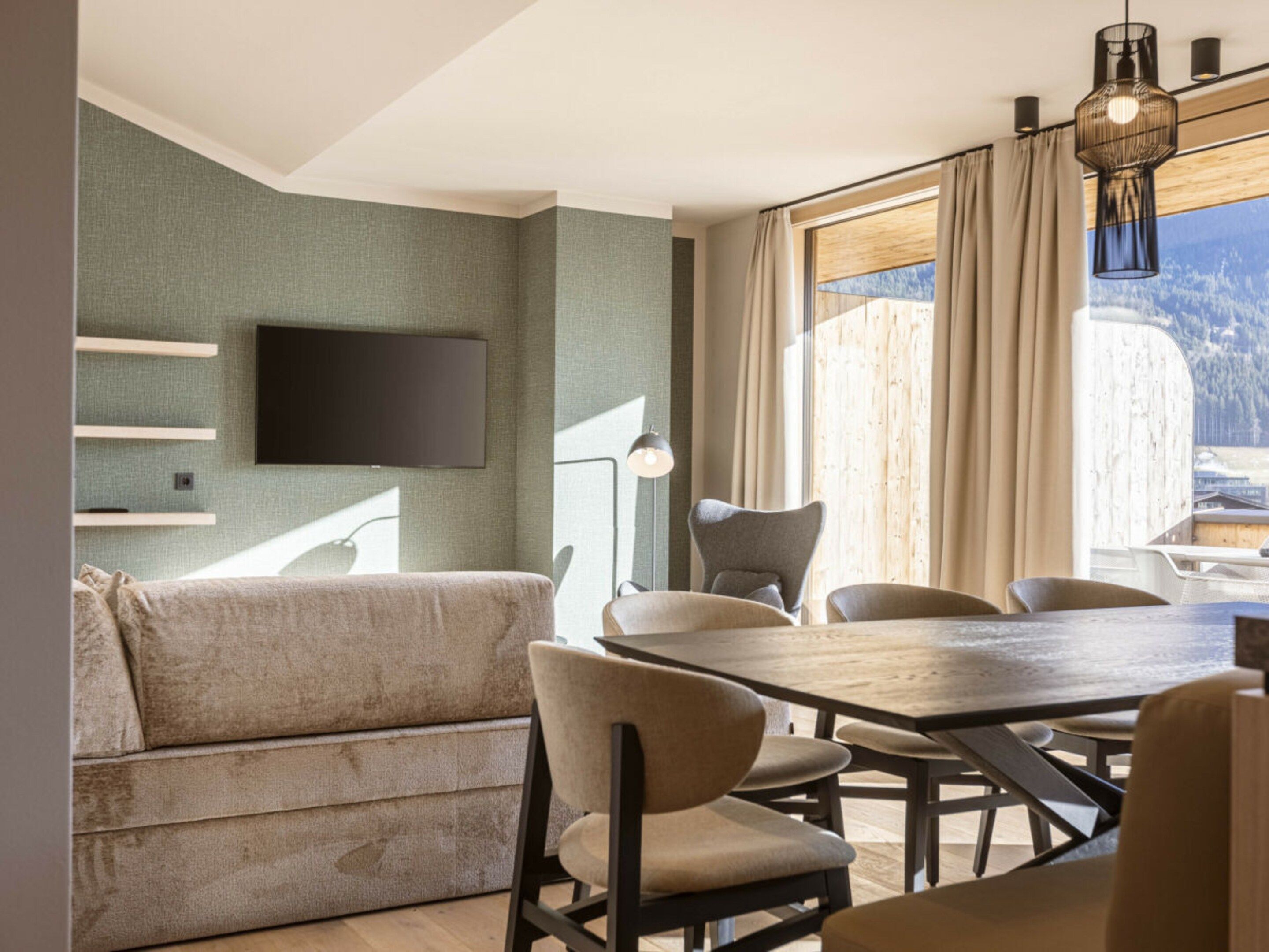 Een moderne eethoek met een houten tafel en stoelen, een flatscreen televisie aan de muur en uitzicht op een bergachtig landschap door grote ramen.