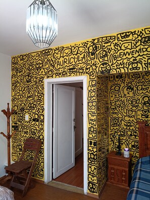 Quarto com parede amarela estampada, um cabideiro de madeira, uma cadeira de madeira, uma cama com cobertor azul e uma porta branca.